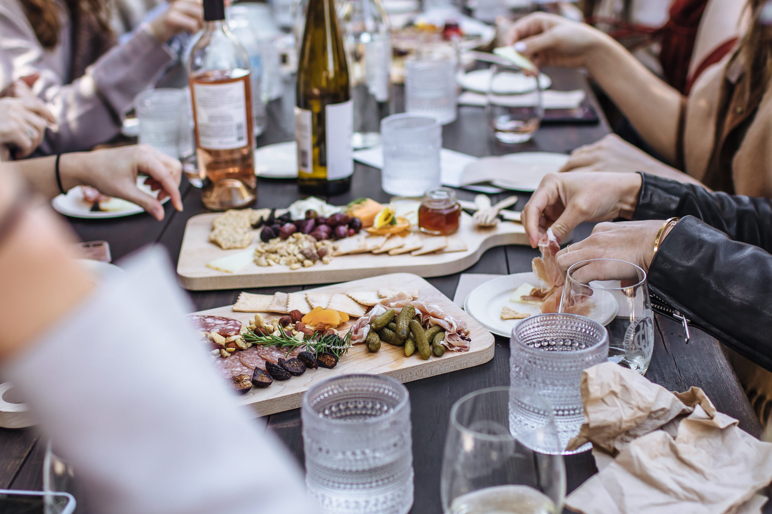 picnic table top filled with food and wine with peoples hands showing around the outside