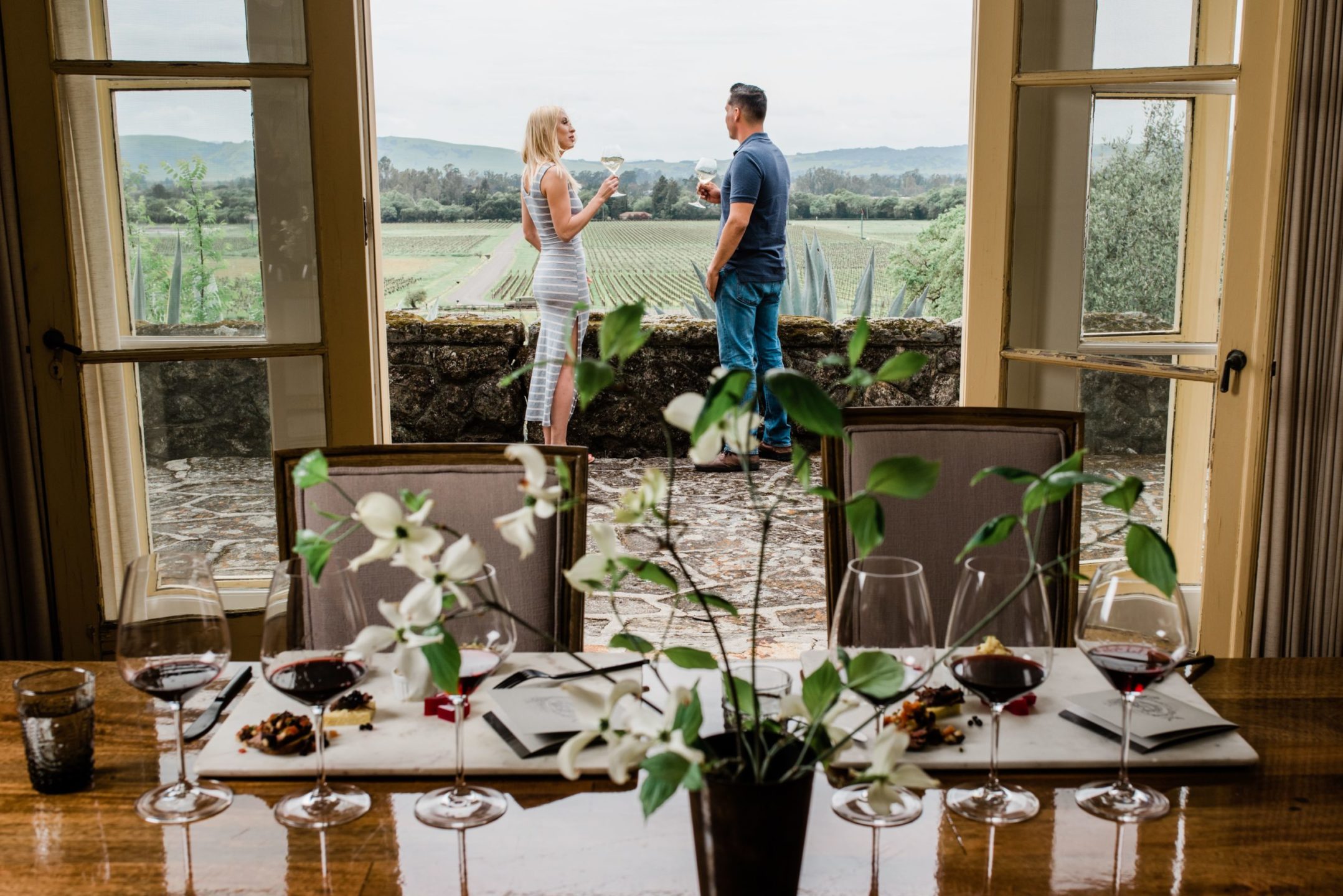 Click to Join the Bacchus Club Image of man and woman on the terrace outside of room having a private Heritage Tasting experience with small plates of food, wine glasses and flowers on the table