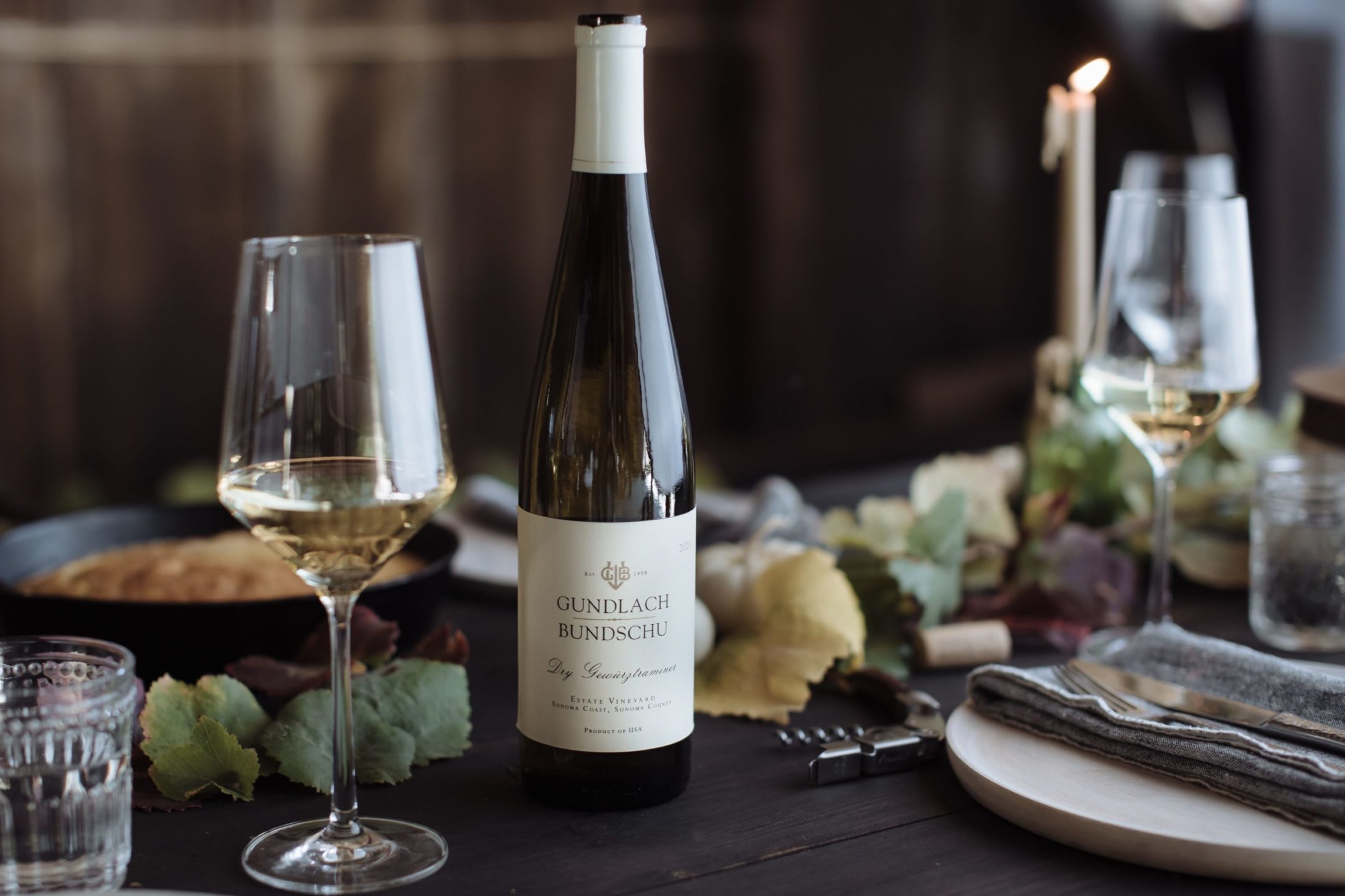 Bottle and two glasses of white wine on a wooden table next to utensils and grape plant leaves