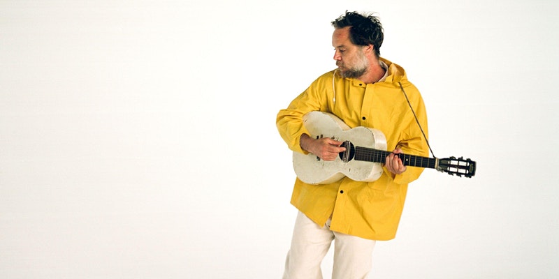 Man with beard in yellow raincoat playing white guitar looking down on a white background
