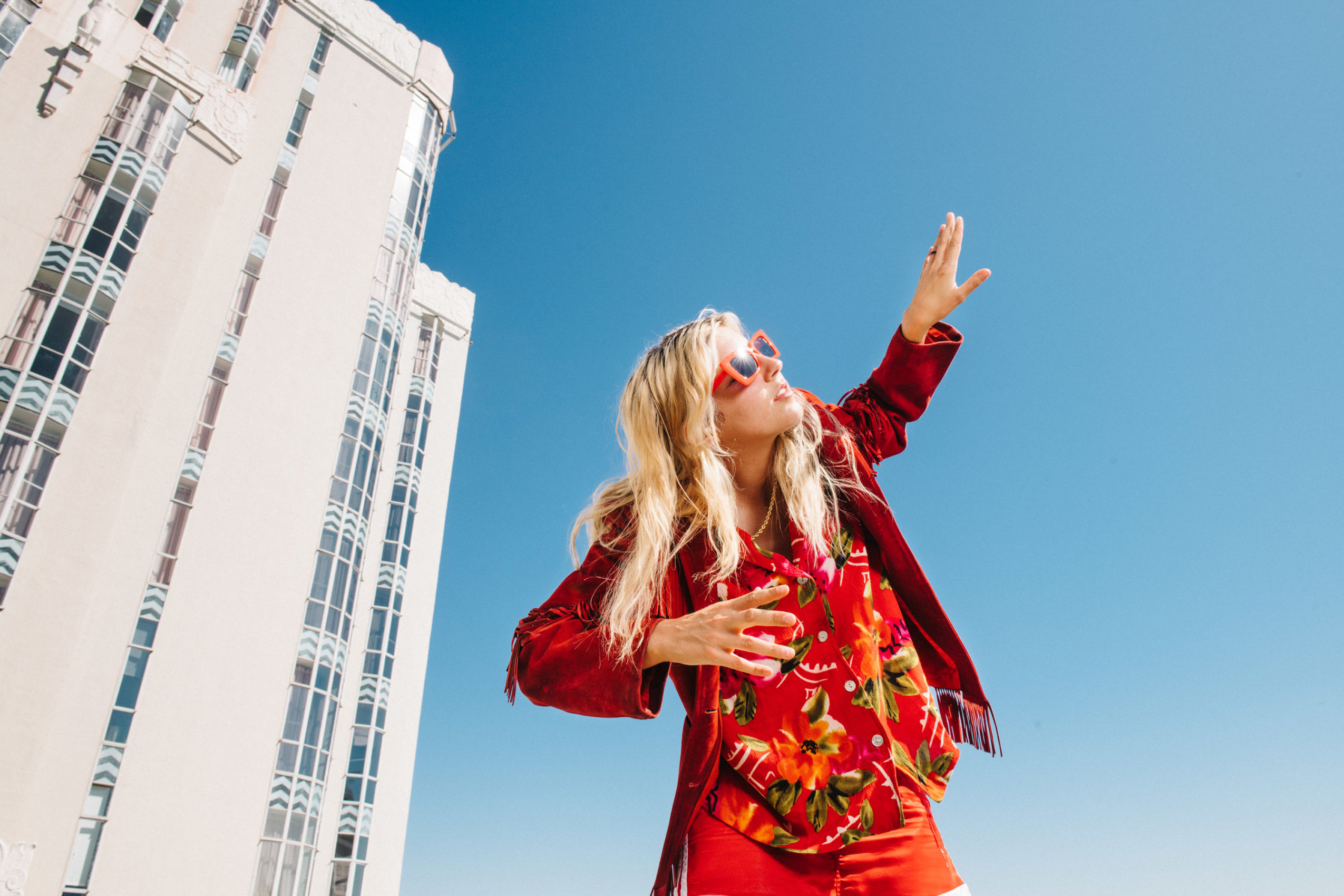 Blonde woman with hand held high in sunglasses and red clothing with a building in the background