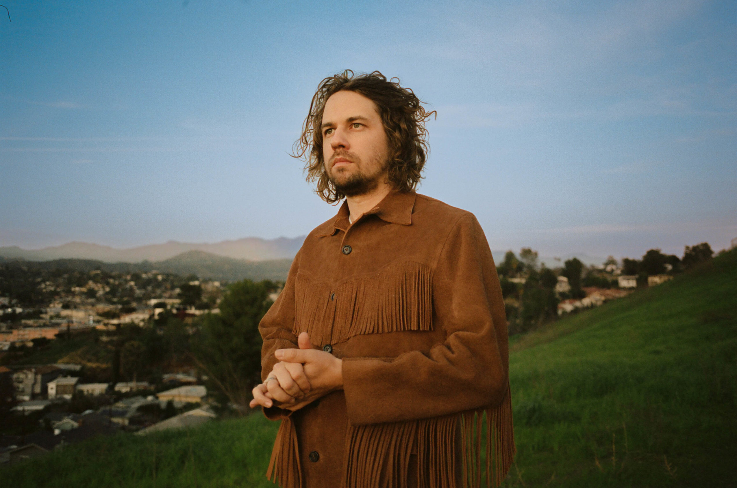 Dark Haired man in suede jacket in green field with blue sky