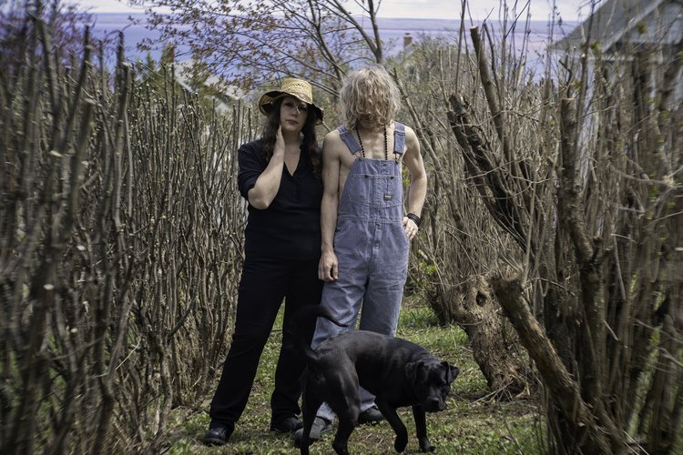 two people in a field branch only trees one person in overalls with another person in black whirt and pants with black dog