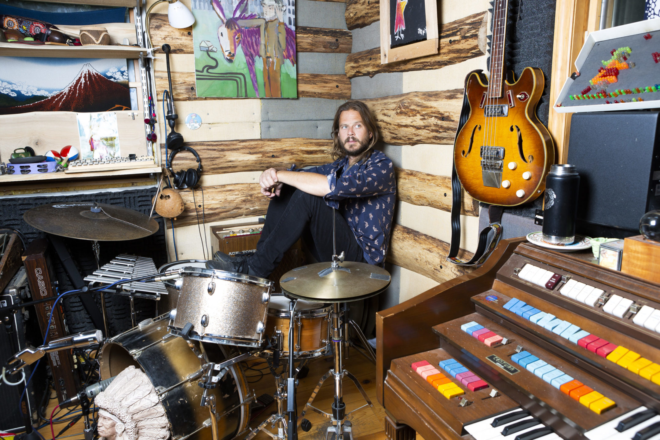 Long-haired person with green eyes sitting behind a drum kit in the corner of a wall next to ancient musical instruments