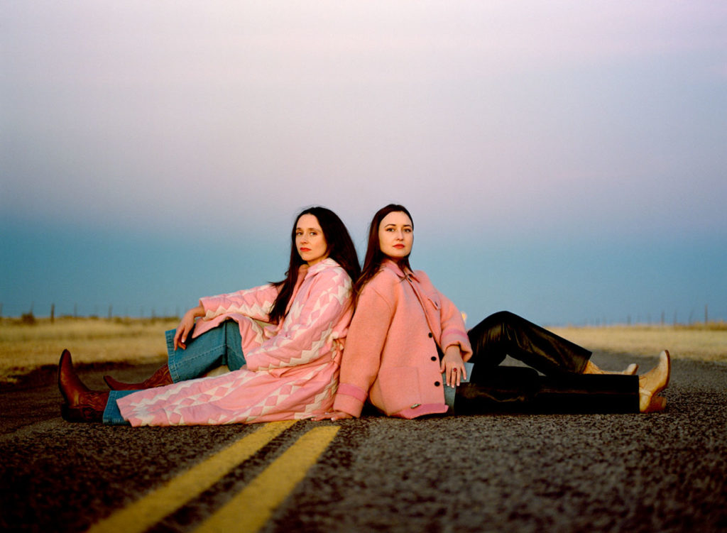 2 women in pink jackets sitting back to back on a road with a double yellow line down the middle
