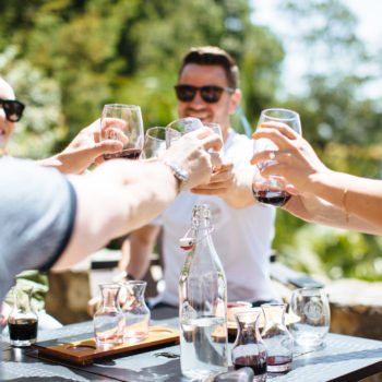 People sitting at a small gray table toasting wine glasses with blurry trees in the background
