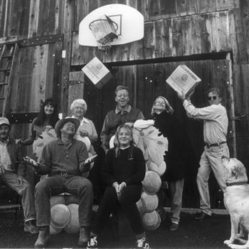 Black and white photo of people sitting and standing in front of a wood barn