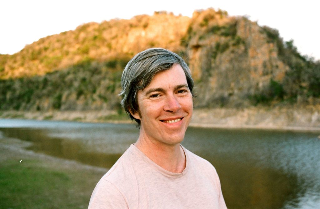 Bill Callahan standing in front of a lake with a hill behind