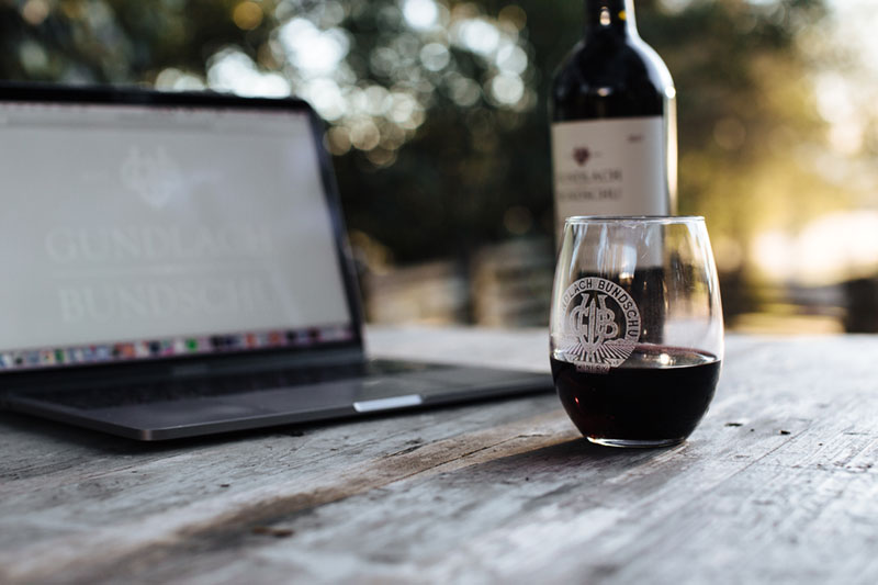 Glass of red wine in a stemless glass on a wood table with an open laptop in the background