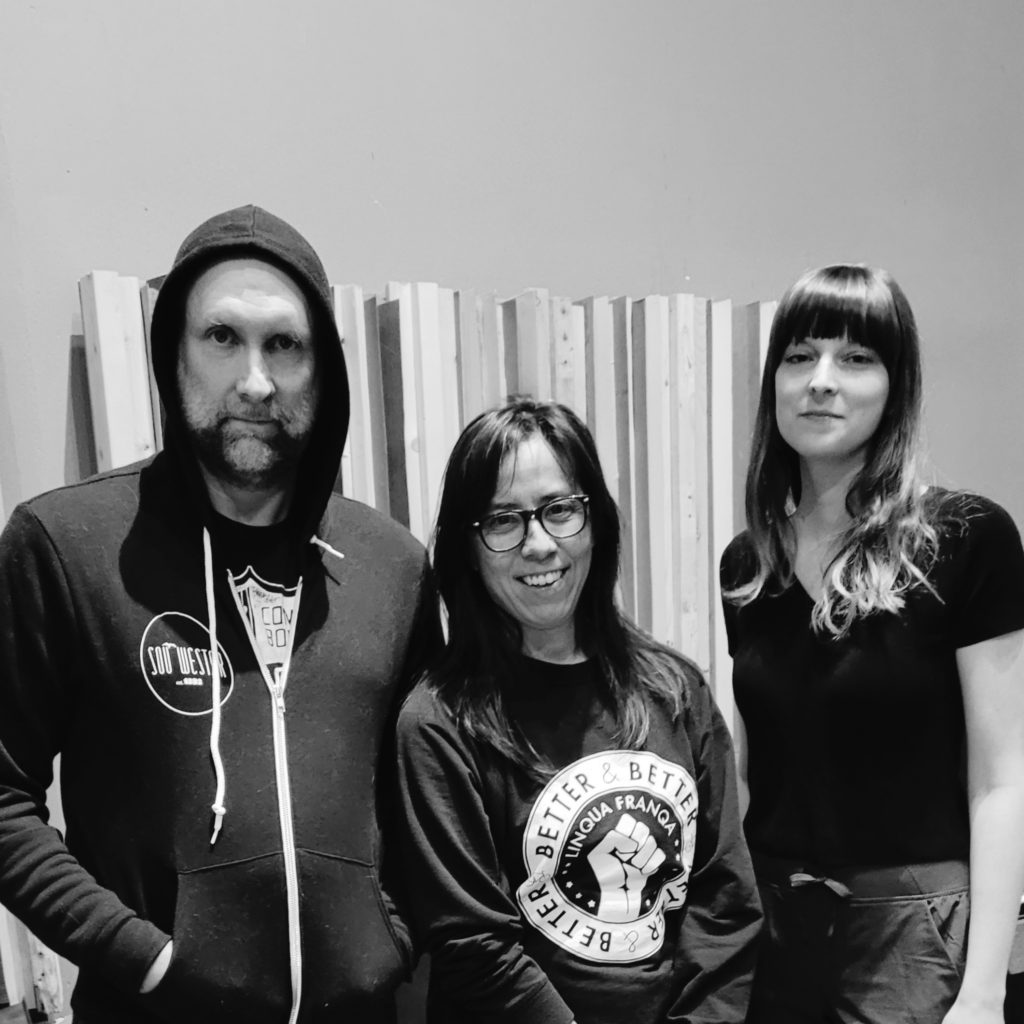 Black and white image of 3 people standing with boards leaning on the wall in the background