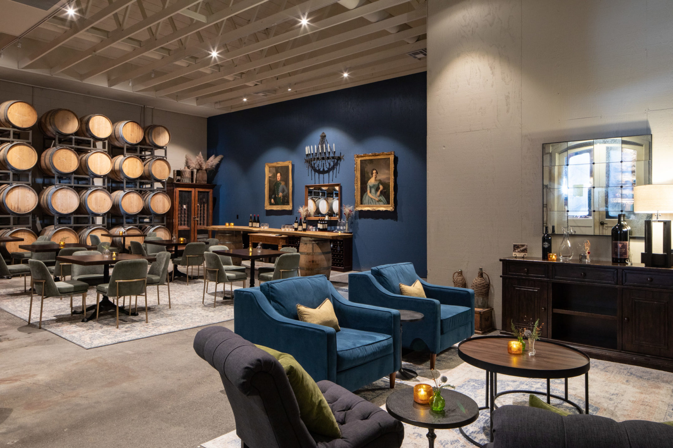 A room with beige and blue walls, two blow chairs with two beige pillows, a two-tier table and two portraits on the blue wall with two people at a barrel table in the background.